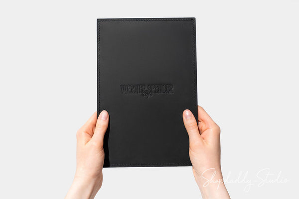 A person's hands holding a black leather journal or notebook cover with 'Wanderlust' embossed on the front, set against a plain light gray background.