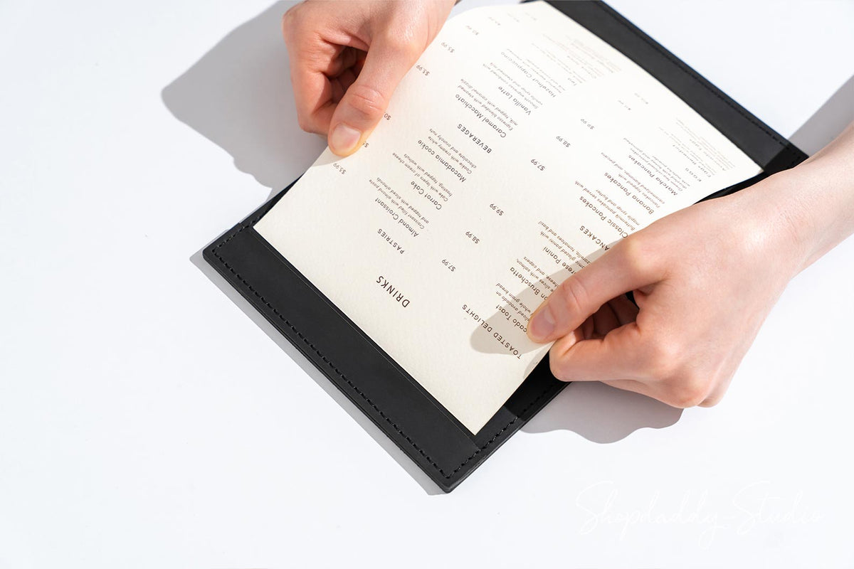 A black faux-leather menu folder with a cream-colored paper menu insert being held open by two hands against a clean white background.