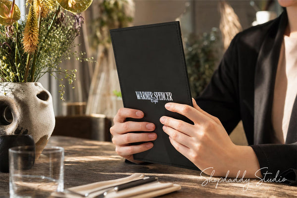 A person in a black blazer holds a black leather-bound book or menu with the title 'Warner Speakr' visible on the cover, set in a warm, sunlit indoor environment with plants and a table setting.