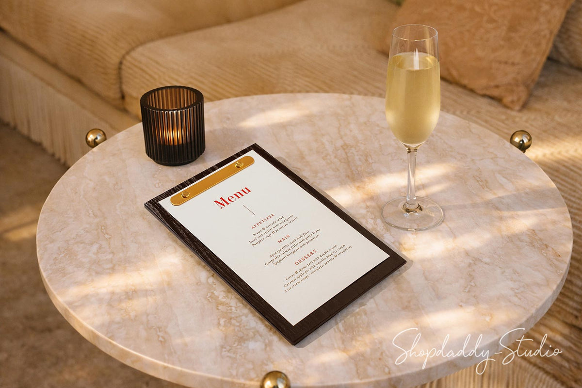 A black-framed menu with a gold accent band and white page sits on a round light marble coffee table beside a lit black candle and a glass of champagne, with a beige sofa in the background.