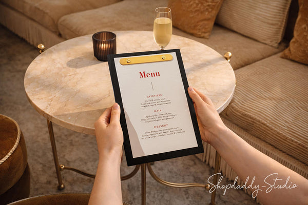 A person holds a black-framed printed menu in a warm indoor setting, with a round marble coffee table, cushions, and a glass of champagne in the background.