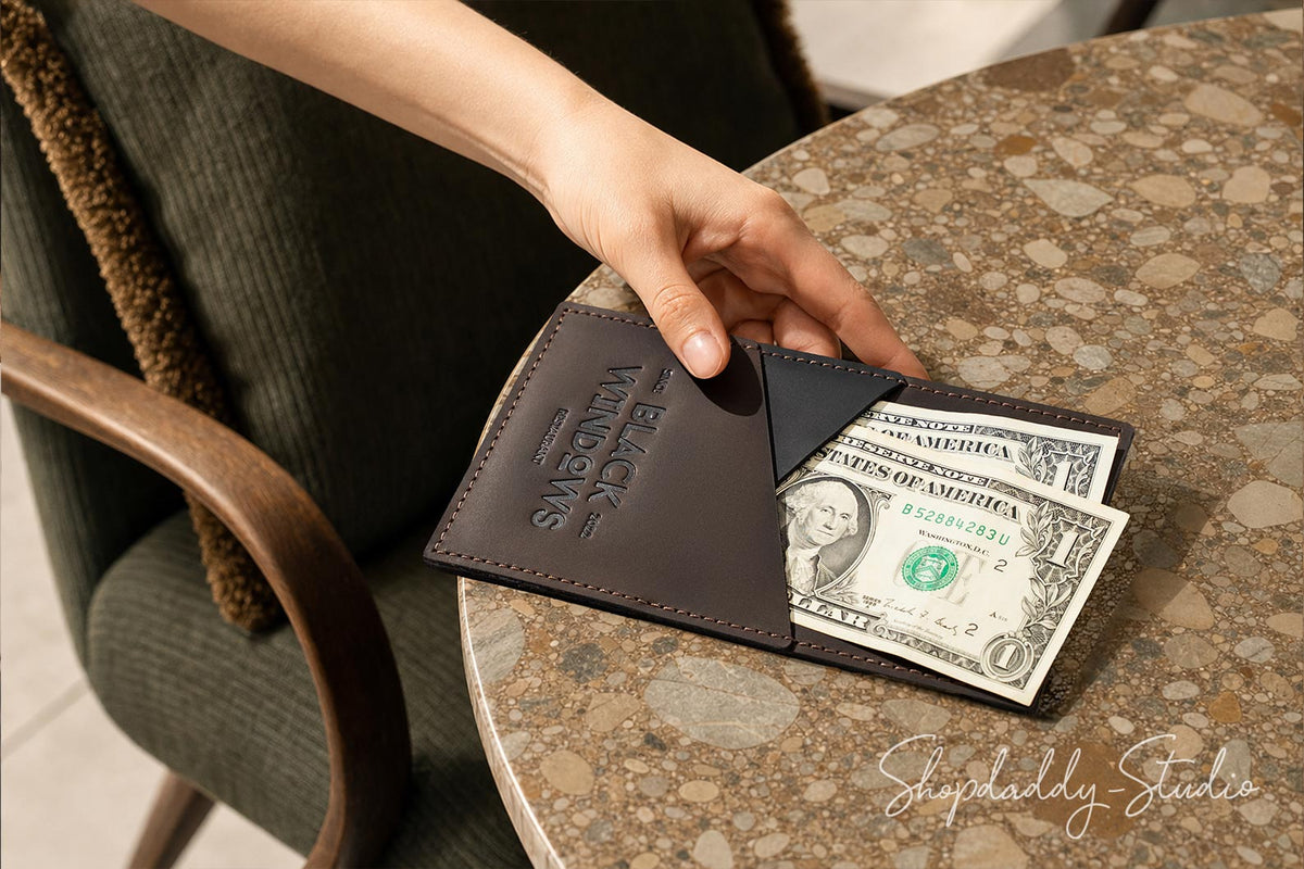 A hand holding an open dark brown leather wallet with the brand name 'BLACK VAMBOIS' embossed on the front, with two US dollar bills partially visible, set on a round terrazzo table next to a chair.