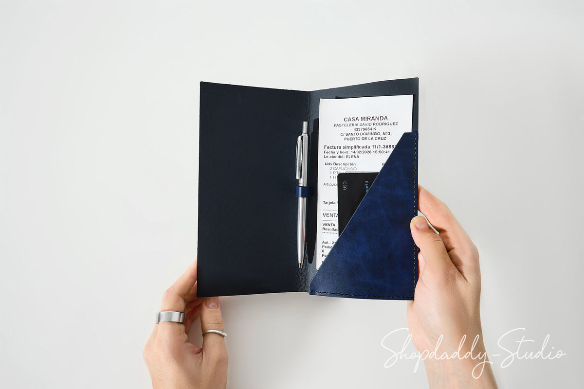A person's hands holding open a navy blue passport cover or organizer, showing a white insert page, a silver pen, and a black phone inside. The photo is taken against a clean white background with a Shopdaddy Studio watermark in the corner.