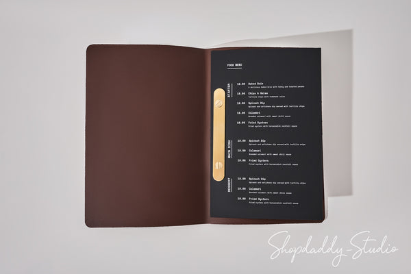 An open menu booklet or folder on a light gray background. The left page is a solid dark brown cover, and the right page is black with white text listing items and a vertical gold accent strip.