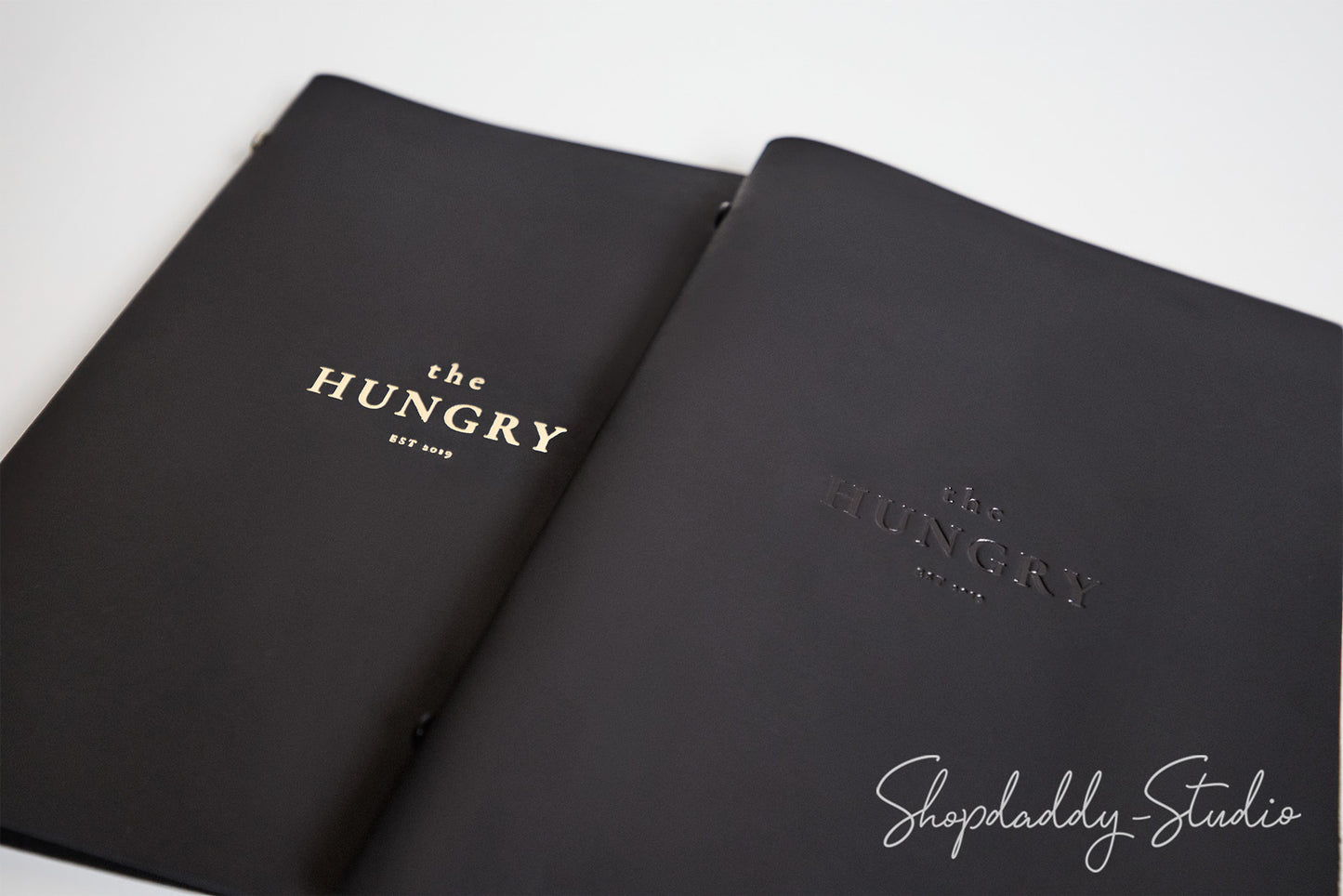 A black hardcover menu or booklet from 'the HUNGRY EST. 2015' shown open on a white background, with gold foil branding on the left cover and embossed branding on the right cover.