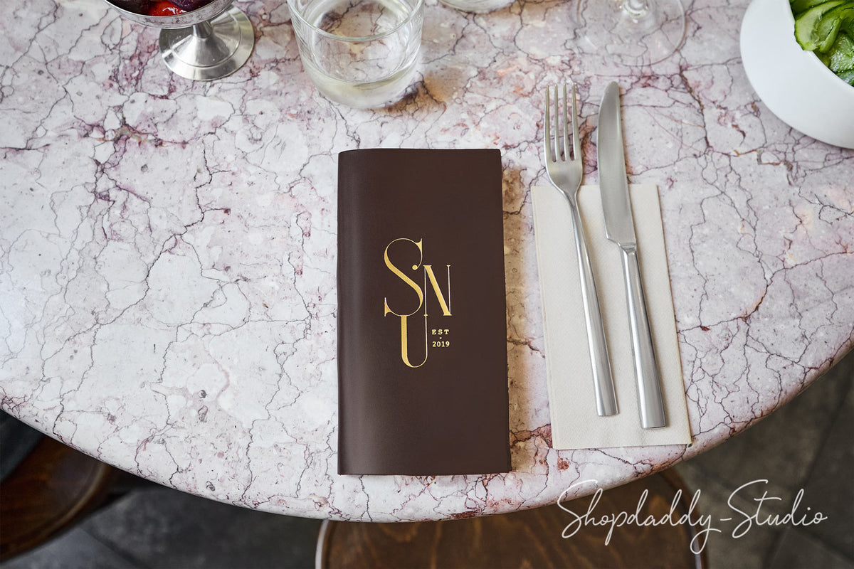 A dark brown hardcover menu or book with gold lettering reading 'SNU' and 'EST. 2019' placed on a round white-and-pink marble table, next to silver cutlery on a cream napkin.