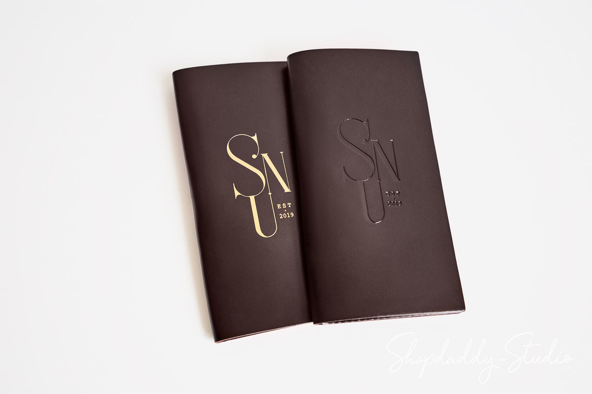 Two dark brown leather-bound notebooks with gold foil 'SNU EST 2025' monogram covers, displayed open on a clean white background.