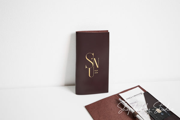 A dark brown leather-bound notebook with gold foil 'SN' lettering stands upright on a white surface, next to a matching brown book and a pen on a white background.