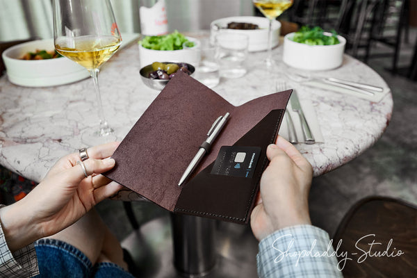 A person holds an open dark brown leather card holder and pen pouch over a round marble table at a restaurant. The pouch contains a black card and a silver pen, with wine glasses, salads, and plates blurred in the background.
