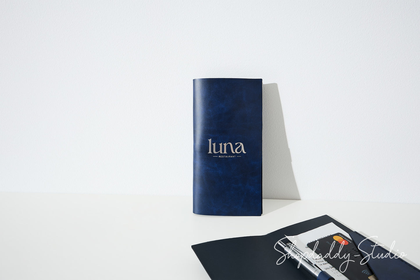 A dark blue textured notebook with the brand name 'luna' printed in white on the front, standing upright on a white surface against a white background. In the foreground, a black notebook and pen are partially visible.