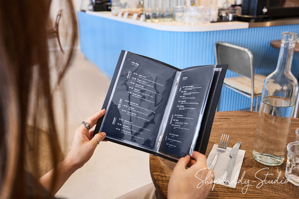 A person in a cafe holding an open black plastic menu binder, viewed from behind. The cafe has a bright blue ribbed wall in the background, a wooden table with a glass bottle of water, and a silver fork and knife on a napkin.