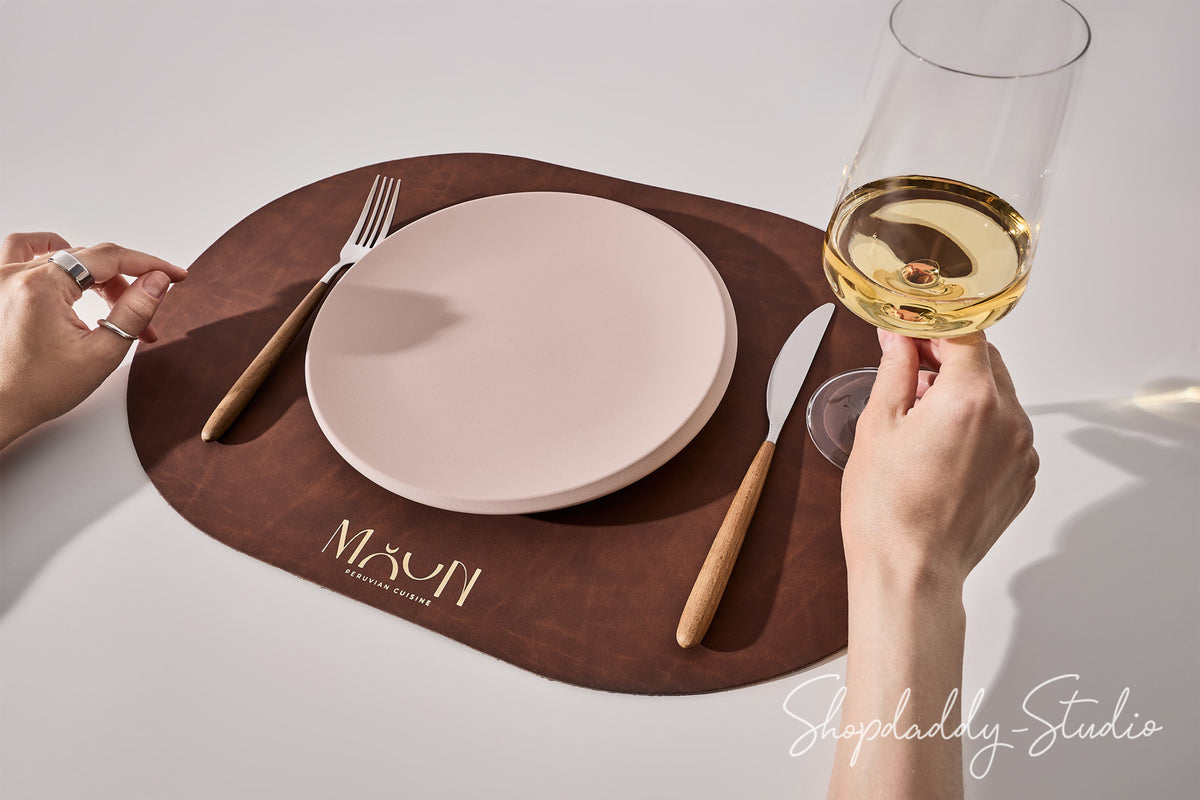 A brown oval placemat with the brand name 'Mxun' printed in gold near the bottom, styled with a white plate, wooden-handled cutlery, and a hand holding a glass of white wine on a white table.