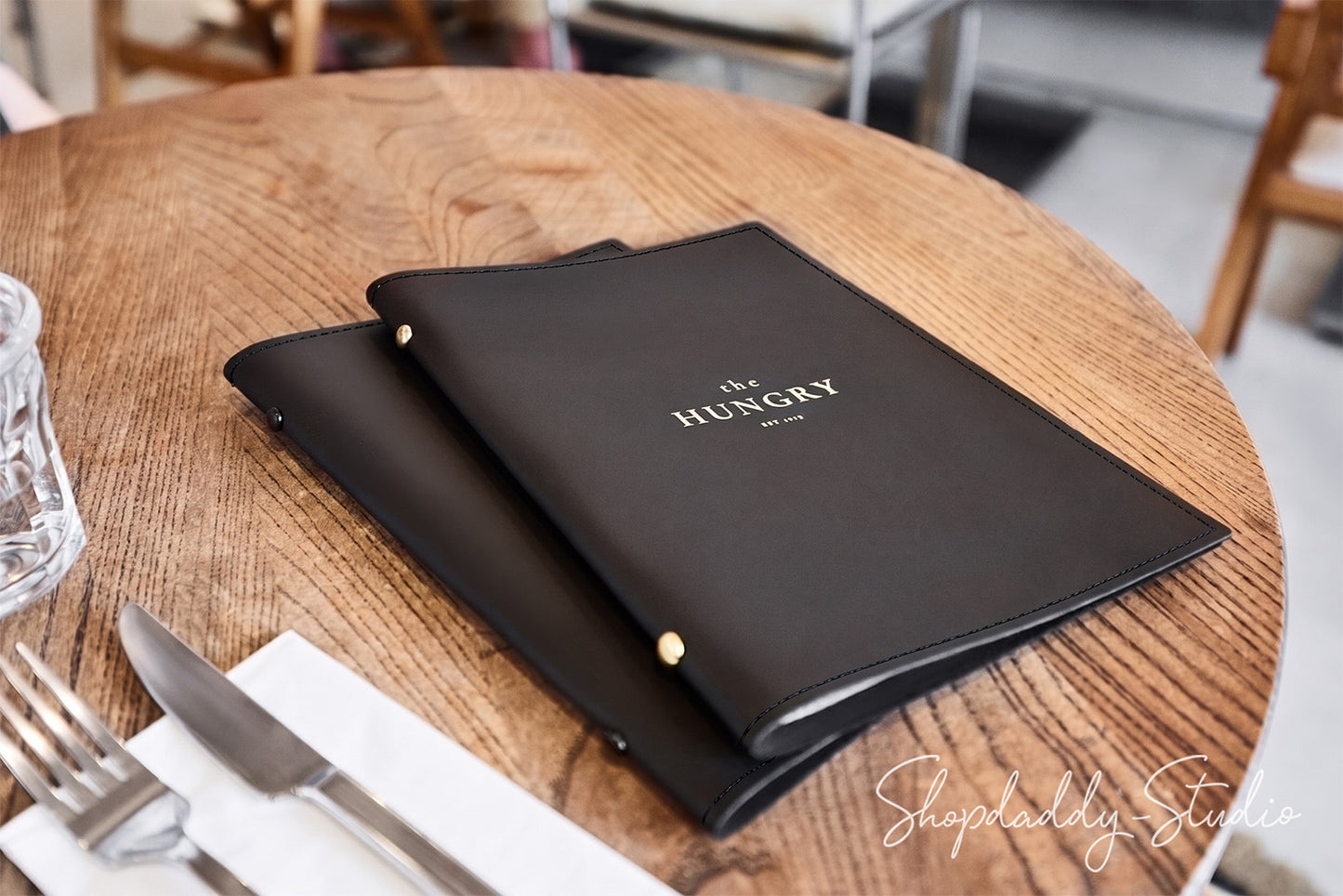 Two black leather menu folders with gold lettering reading 'THE HUNGRY' resting on a round wooden table, with a fork, knife, and glass partially visible in the foreground.