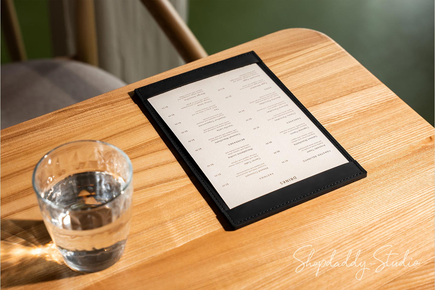 A black leather-bound paper menu pad with printed text lies on a light wood table next to a clear glass of water. The background is softly blurred, showing part of a chair and wall.