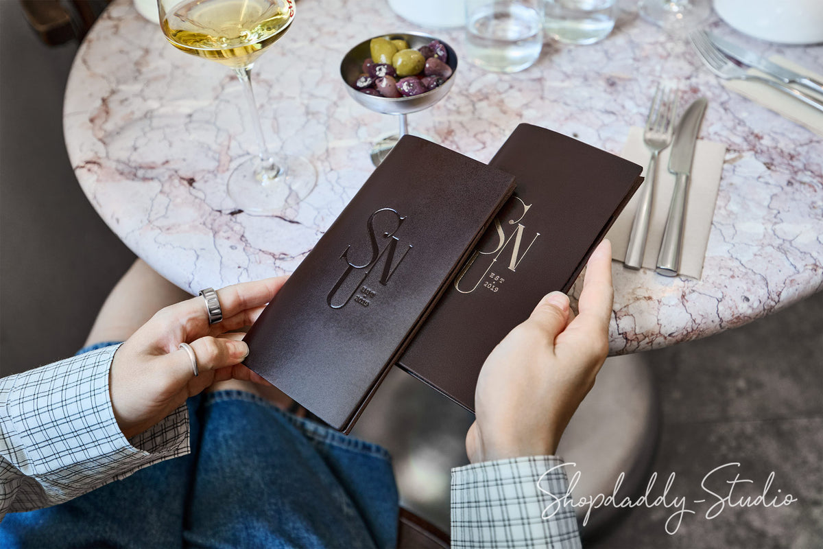 A person holding two dark brown leather menus or booklets with embossed 'SVN' lettering, seated at a round marble table with a wine glass, olives, and cutlery.
