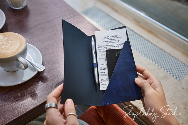 A navy blue leather travel organizer wallet held open by hands at a cafe table, containing a receipt, pen, and passport, with a latte visible in the background.
