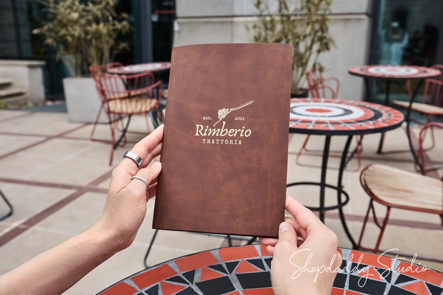A person holds a brown leather-bound menu or booklet with the printed text 'Rimberio Trattoria' and a guitar logo, photographed on an outdoor patio with tables and chairs in the background.