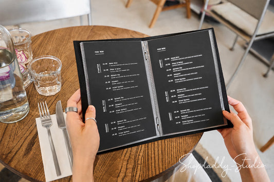 A person holding an open black printed menu at a round wooden table in a cafe setting, with a glass bottle, water glasses, a fork, and knife nearby.
