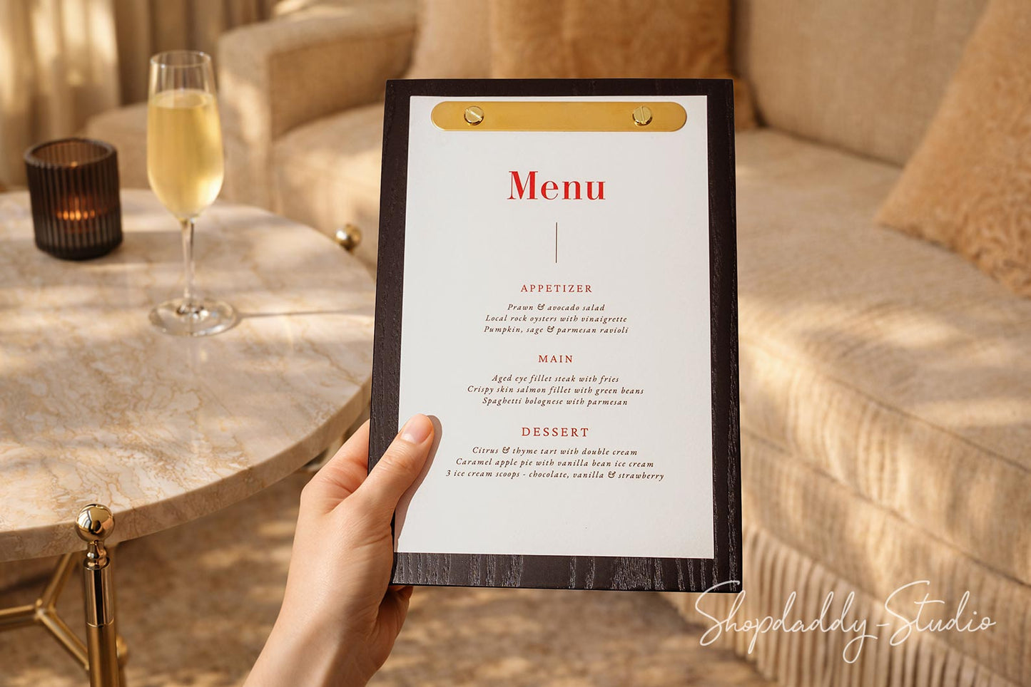 A hand holds a black clipboard-style menu with a gold clip and a white page featuring red text, set against a warm, cozy living room background with a sofa, table, and a glass of champagne.