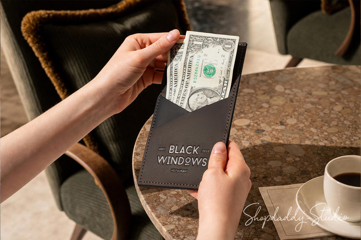 A person's hands holding a dark brown leather wallet with the text 'BLACK WINDOWS' and inserting a one hundred dollar bill, set on a marble table next to a cup of coffee.