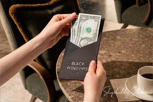A person's hands holding a dark brown leather wallet with the text 'BLACK WINDOWS' and inserting a one hundred dollar bill, set on a marble table next to a cup of coffee.