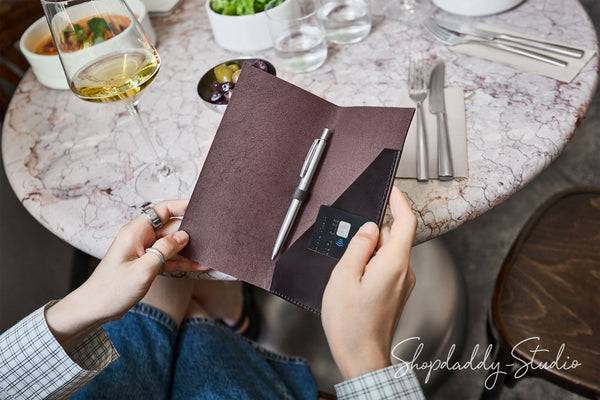 A person holds an open dark brown leather notebook cover at a round marble table in a restaurant setting. The cover contains a pen and a card holder pocket.