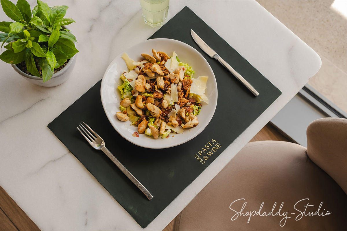 A top-down view of a pasta salad served in a white bowl on a black placemat on a white marble table. The placemat has gold lettering that reads 'PASTA & PINE' and is accompanied by a silver fork and knife.