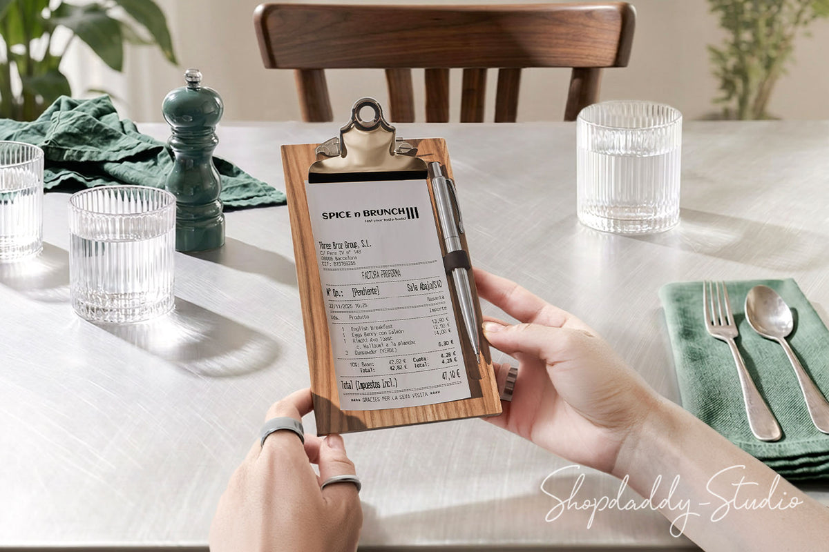 A person holds a wooden clipboard check holder with a printed receipt in a dining room setting. The scene includes a white tablecloth, glassware, a green napkin with silverware, a dark green pepper mill, and a wooden chair in the background.