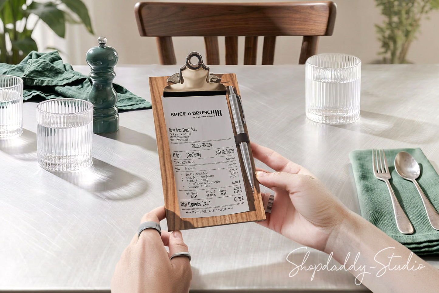 A person holds a wooden clipboard check holder with a printed receipt in a dining room setting. The scene includes a white tablecloth, glassware, a green napkin with silverware, a dark green pepper mill, and a wooden chair in the background.