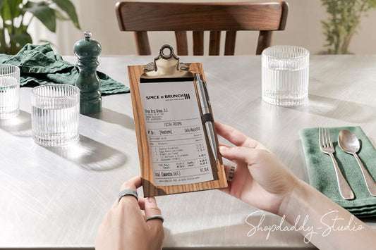 A person holds a wooden clipboard check holder with a printed receipt in a dining room setting. The scene includes a white tablecloth, glassware, a green napkin with silverware, a dark green pepper mill, and a wooden chair in the background.