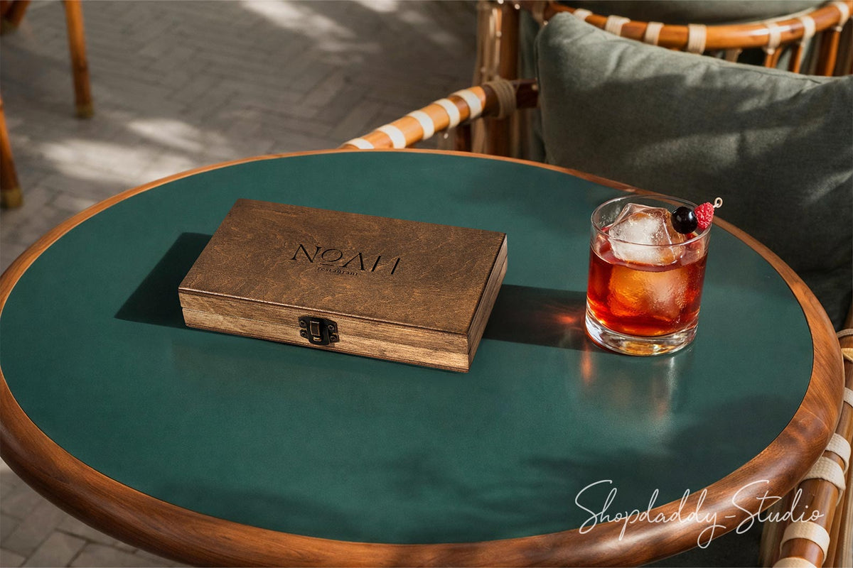 A personalized wooden сheck box engraved with the name 'NOAH' sits on a round green and wood outdoor patio table next to a glass of amber cocktail with an olive garnish.