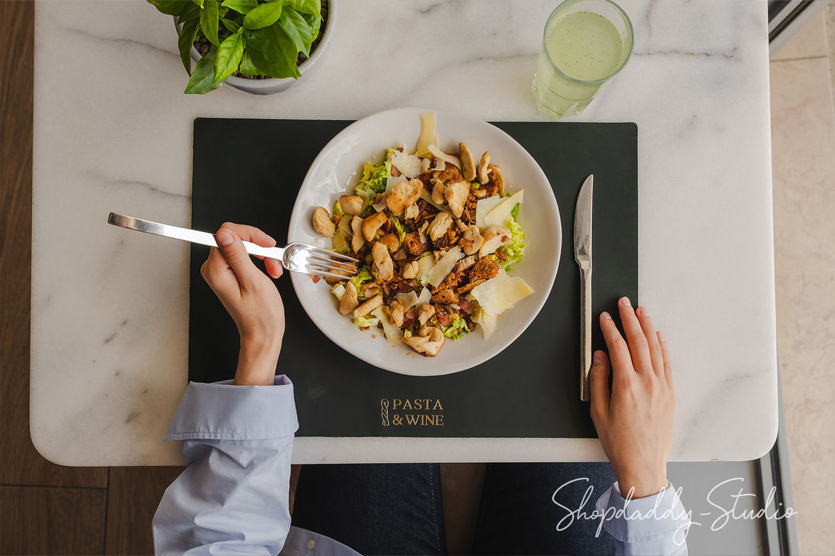 Overhead view of a person eating a pasta salad with chicken and cheese on a white plate, placed on a black placemat with the words 'PASTA & WINE' printed on it. The person's hands and arms are visible, along with a fork, knife, marble table, a glass of water, and a potted basil plant.
