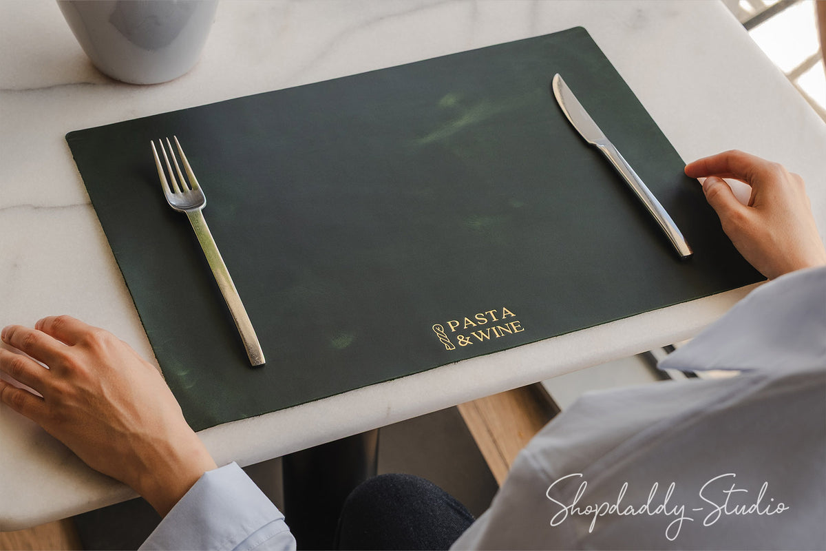 A dark green rectangular placemat on a white marble table, featuring the gold foil text 'PASTA & WINE'. A silver fork and knife are placed on the placemat, with a person's hands and arms resting near it, wearing a light blue shirt.
