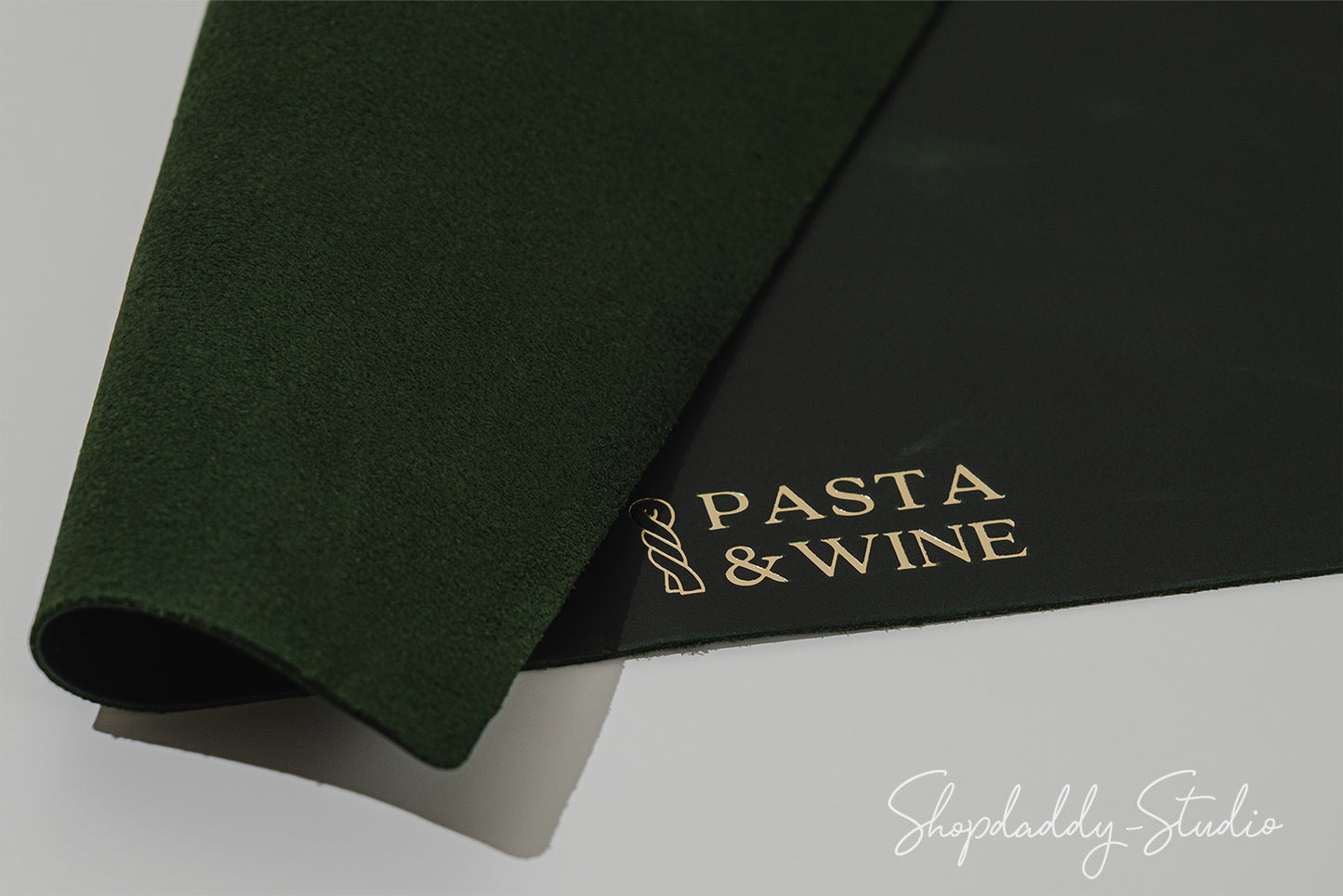 A rolled dark green felt or suede-like mat partially covering a black rectangular card printed with the words 'PASTA & WINE' in gold, photographed on a light gray background.