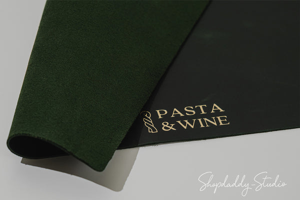 A rolled dark green felt or suede-like mat partially covering a black rectangular card printed with the words 'PASTA & WINE' in gold, photographed on a light gray background.