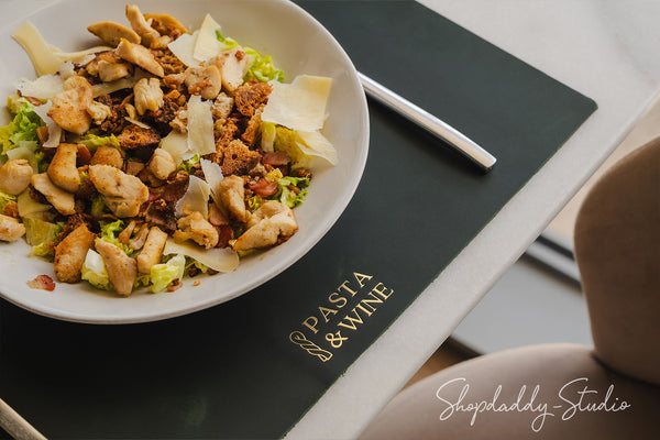 A close-up shot of a fresh Caesar salad in a white bowl, featuring chicken, crumbled bacon, parmesan cheese shavings, and lettuce, set on a black placemat with gold lettering that reads 'PASTA & WINE'.