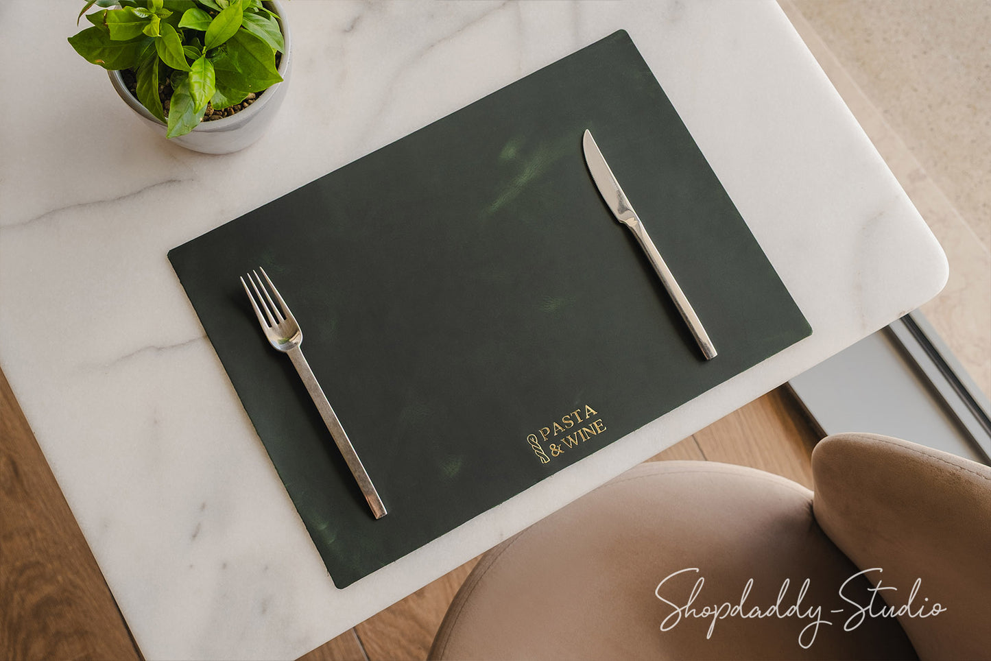 A dark green rectangular placemat on a white marble table, featuring the gold printed text 'Pasta & Wine' and styled with a silver fork and knife. A small potted green plant is visible in the top-left corner.