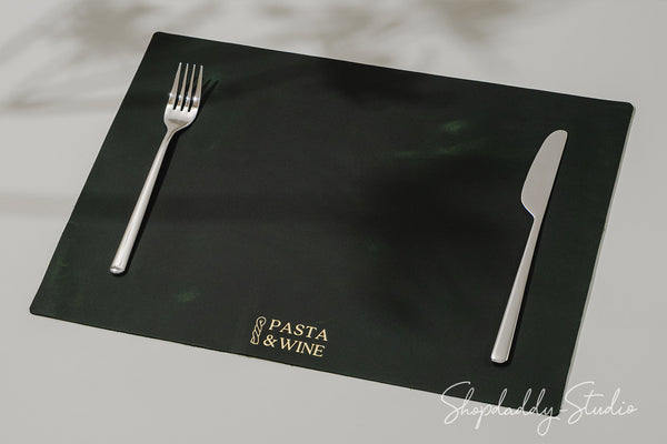 A green rectangular placemat with gold 'Pasta & Wine' branding, styled with a silver fork on the left and knife on the right, photographed on a light gray surface.