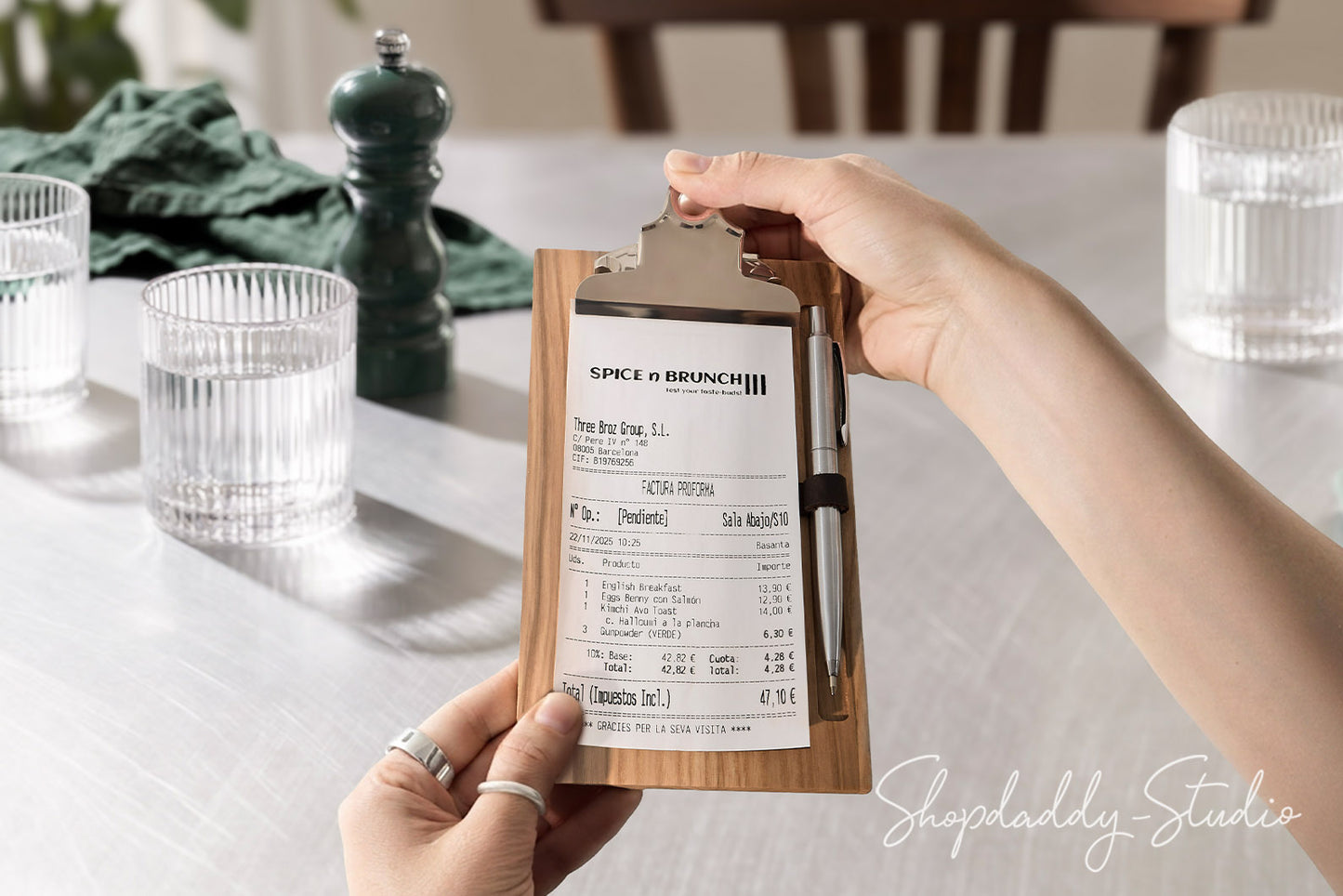A person holds a wooden clipboard with a printed receipt and pen in a restaurant setting. The clipboard has a light wood grain finish and a silver metal clip.