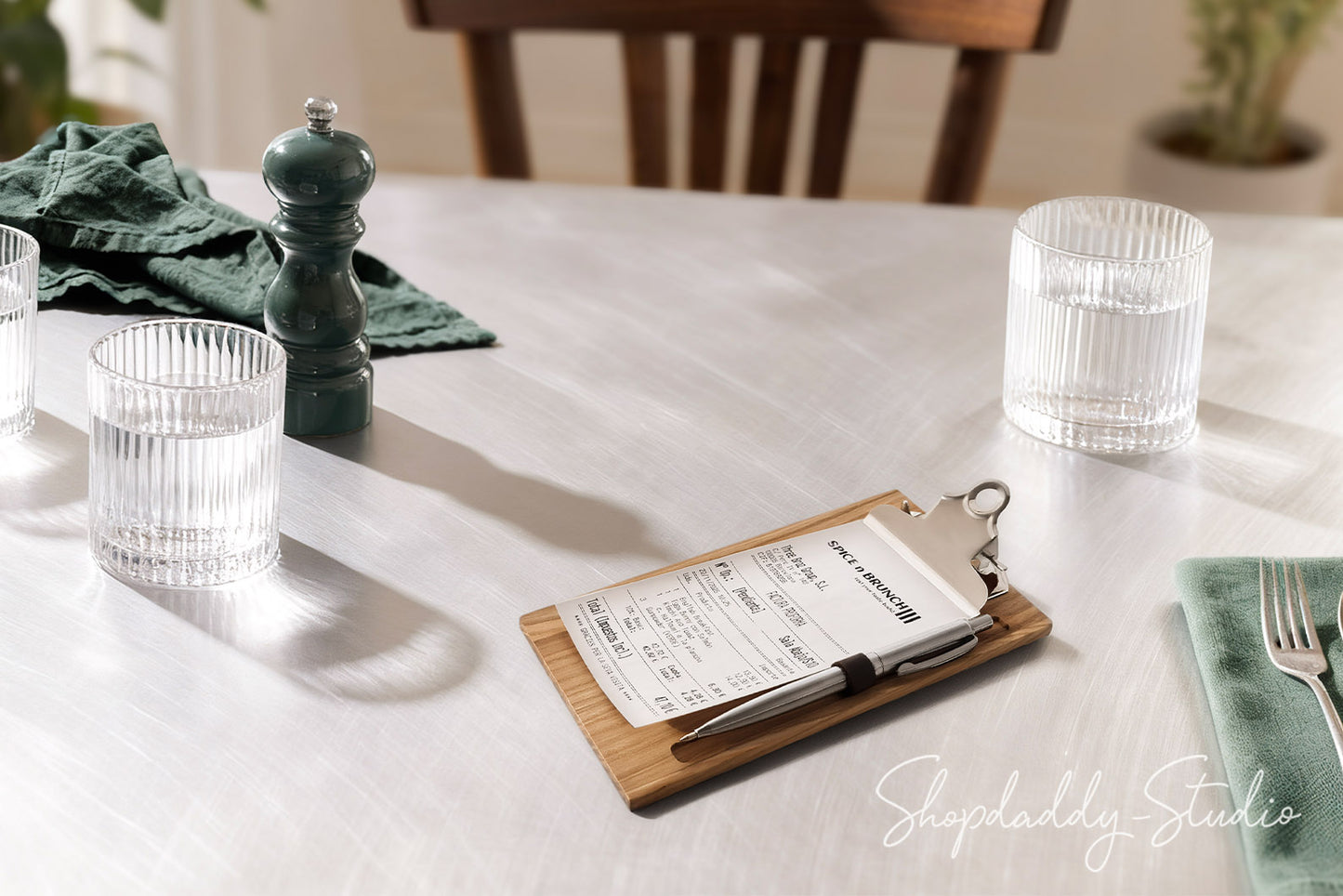 A wooden rectangular board holding a printed receipt or menu card, with a black pen resting on it, set on a white tablecloth in a dining setting. Clear ribbed drinking glasses, a dark green pepper mill, a folded green napkin, and a fork are also visible.