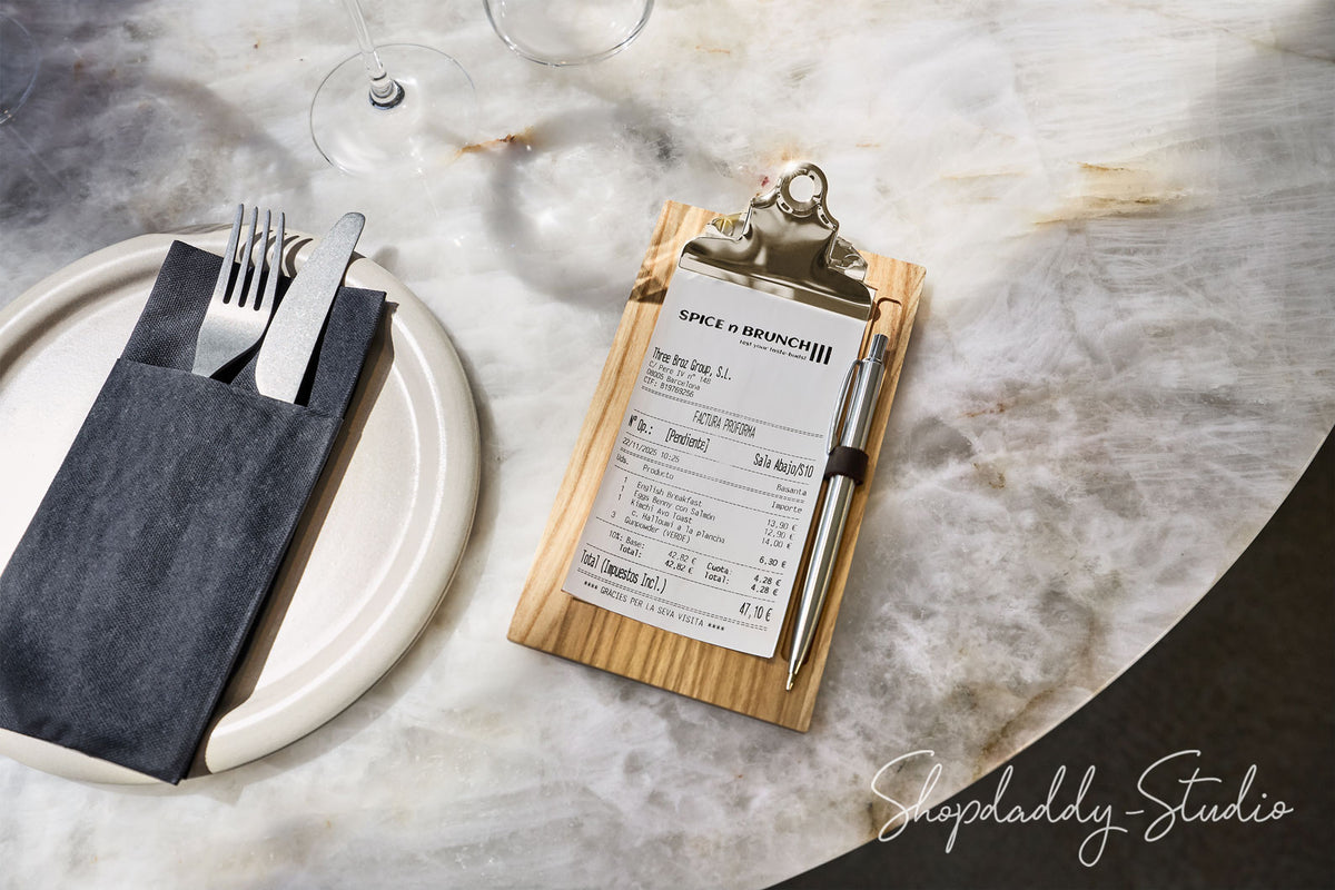 A wooden clipboard holding a printed receipt, styled on a white plate with a dark napkin and silver cutlery on a marble tabletop. A wine glass base is visible in the background.