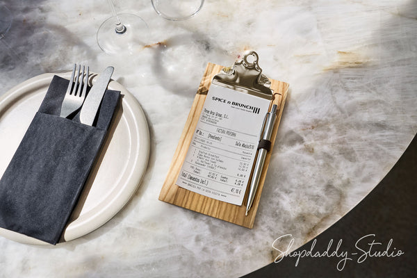 A wooden clipboard holding a printed receipt, styled on a white plate with a dark napkin and silver cutlery on a marble tabletop. A wine glass base is visible in the background.