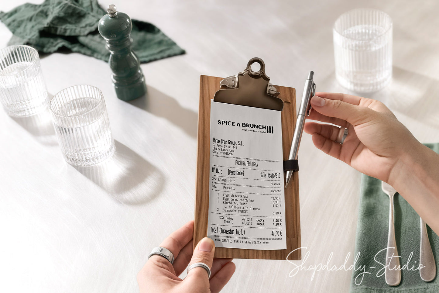 A person holds a clipboard with a restaurant receipt and a pen on a white tabletop, surrounded by glassware, a green napkin, and a pepper mill. The receipt is clipped to the clipboard, and a 'SophieDaddy Studios' watermark is visible in the lower right corner.