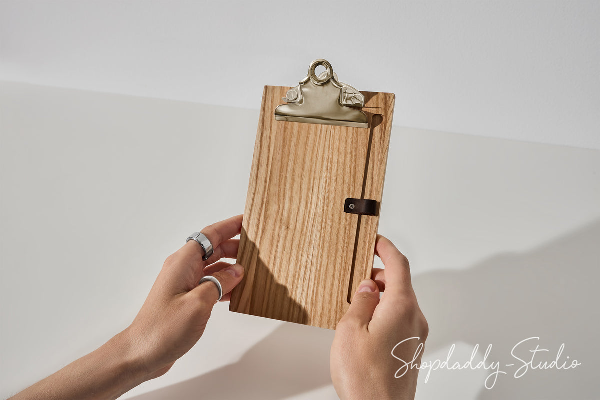 A wooden clipboard with a natural wood grain finish, a silver metal clip, and a small black leather strap closure, held by two hands against a white background.