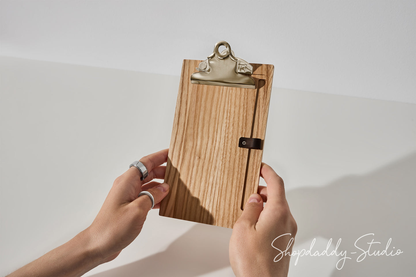 A wooden clipboard with a natural wood grain finish, a silver metal clip, and a small black leather strap closure, held by two hands against a white background.