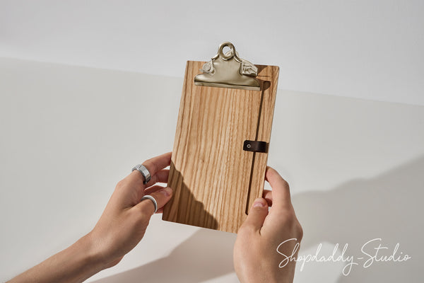 A wooden clipboard with a natural wood grain finish, a silver metal clip, and a small black leather strap closure, held by two hands against a white background.