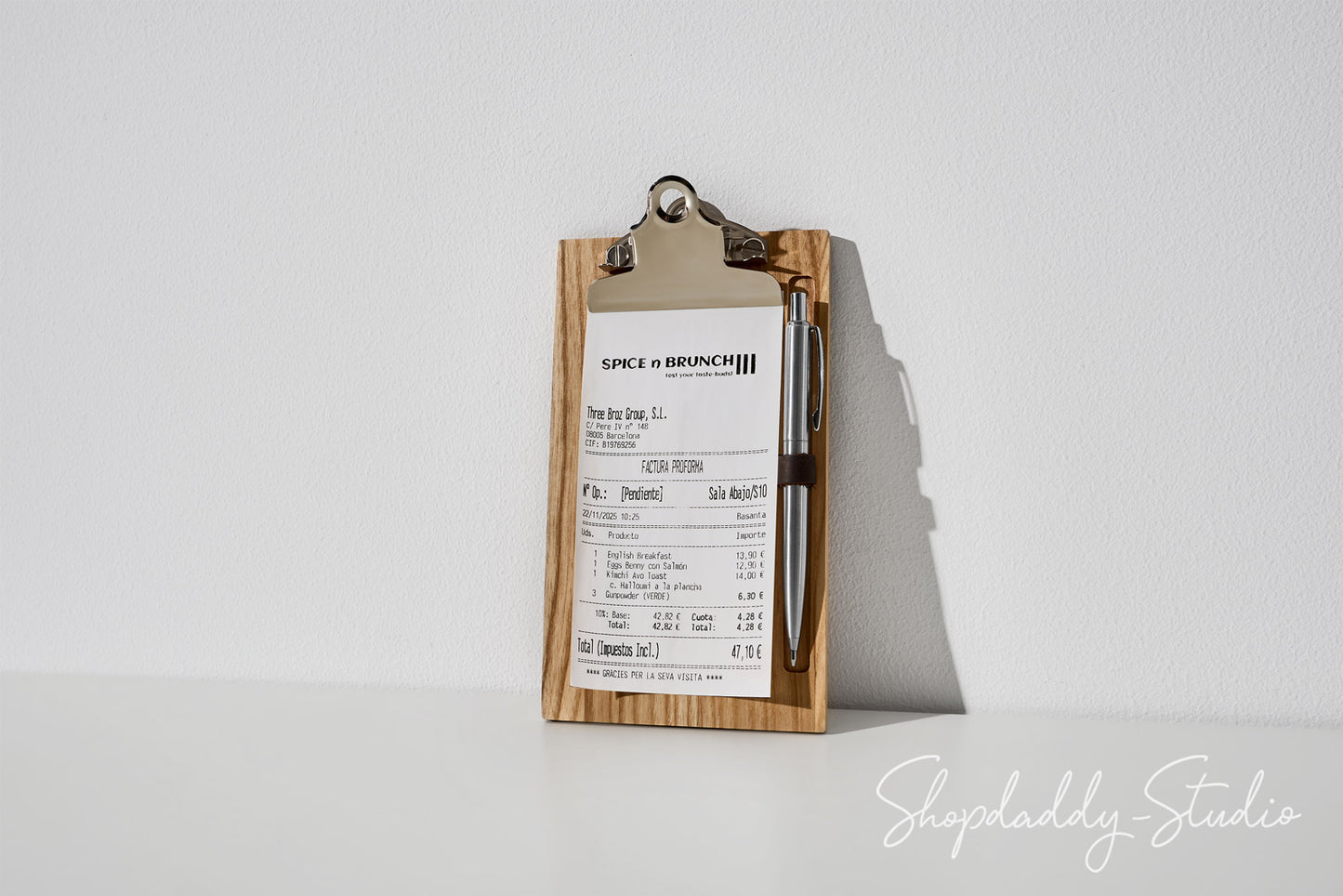 A wooden clipboard with a metal clip holding a printed receipt or ticket and a pen, photographed on a white surface against a white wall with a soft shadow. A Shopdaddy Studio watermark appears in the lower right corner.