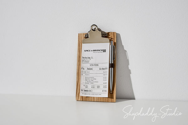 A wooden clipboard with a metal clip holding a printed receipt or ticket and a pen, photographed on a white surface against a white wall with a soft shadow. A Shopdaddy Studio watermark appears in the lower right corner.