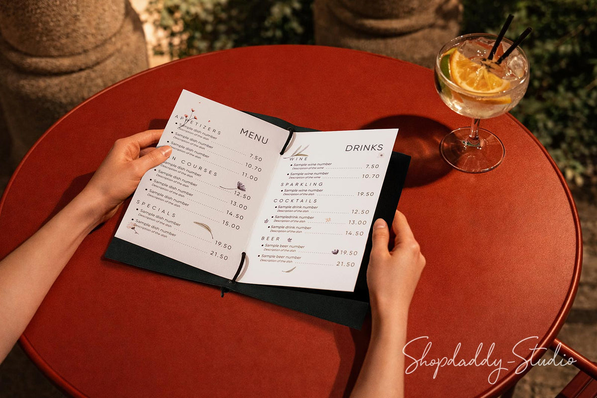 A top-down view of a person holding an open printed menu on a round red table, with a cocktail glass and an orange slice nearby. The scene is photographed from above in a restaurant or cafe setting.