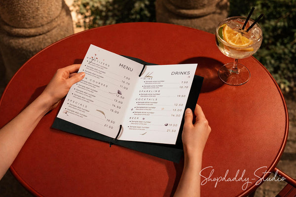 A top-down view of a person holding an open printed menu on a round red table, with a cocktail glass and an orange slice nearby. The scene is photographed from above in a restaurant or cafe setting.
